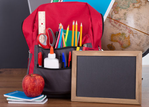 Assortment of school supplies in a bookbag with a blank chalkboard on a wooden desktop